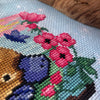 Extreme closeup of righthand side of platypus cross stitch pattern. Platypus is sniffing the pink flowers, which have black cores. Stitches are neat and tidy. Colors are vibrant. Platypus is adorable.