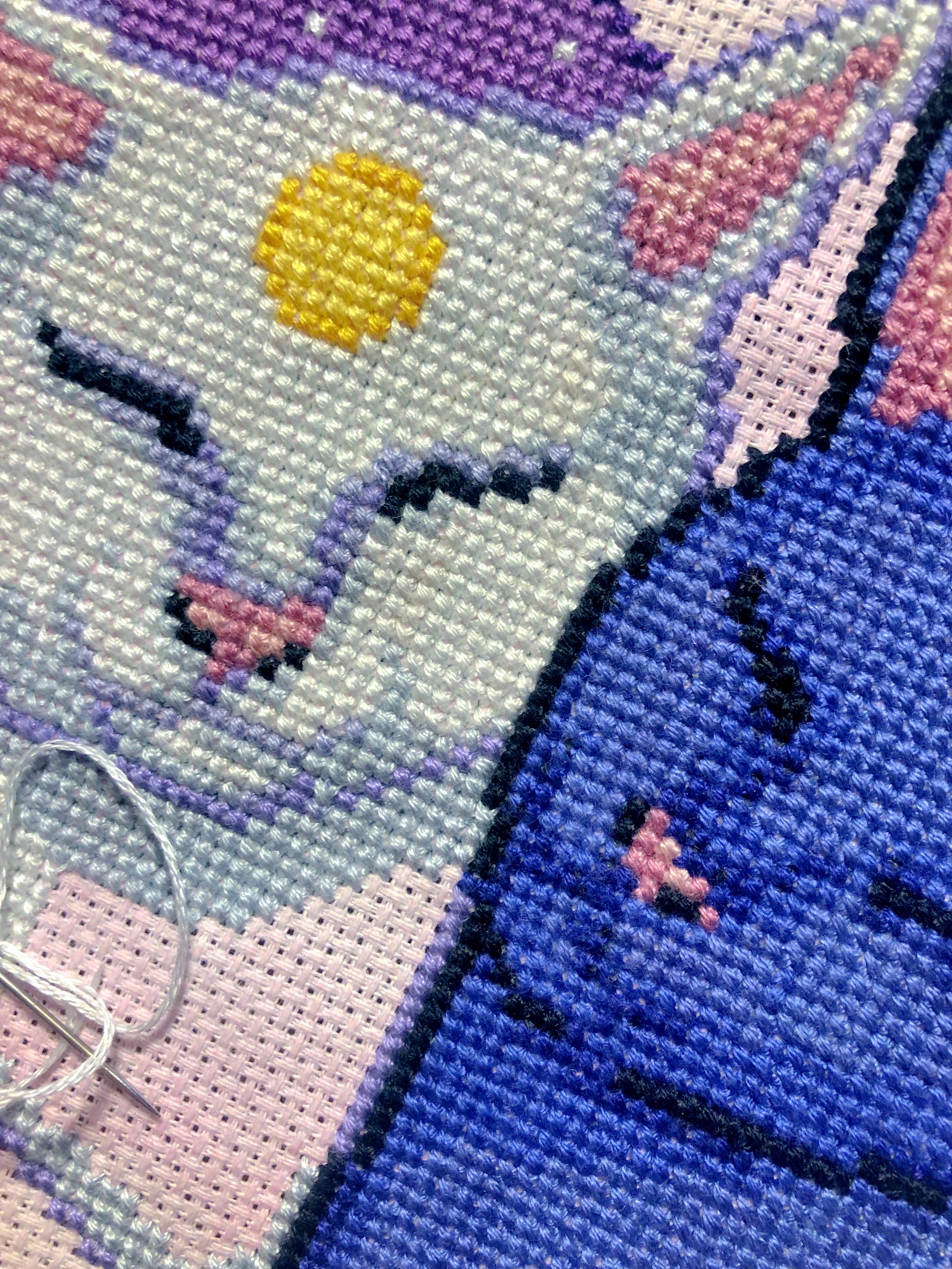 Extreme closeup of Sun and Moon cats cross stitch pattern and picture of the work in progress, with needle and thread displayed. Both cats have their eyes closed. Stitches are neat and tidy. Shading is done in complementary colors like lilac.