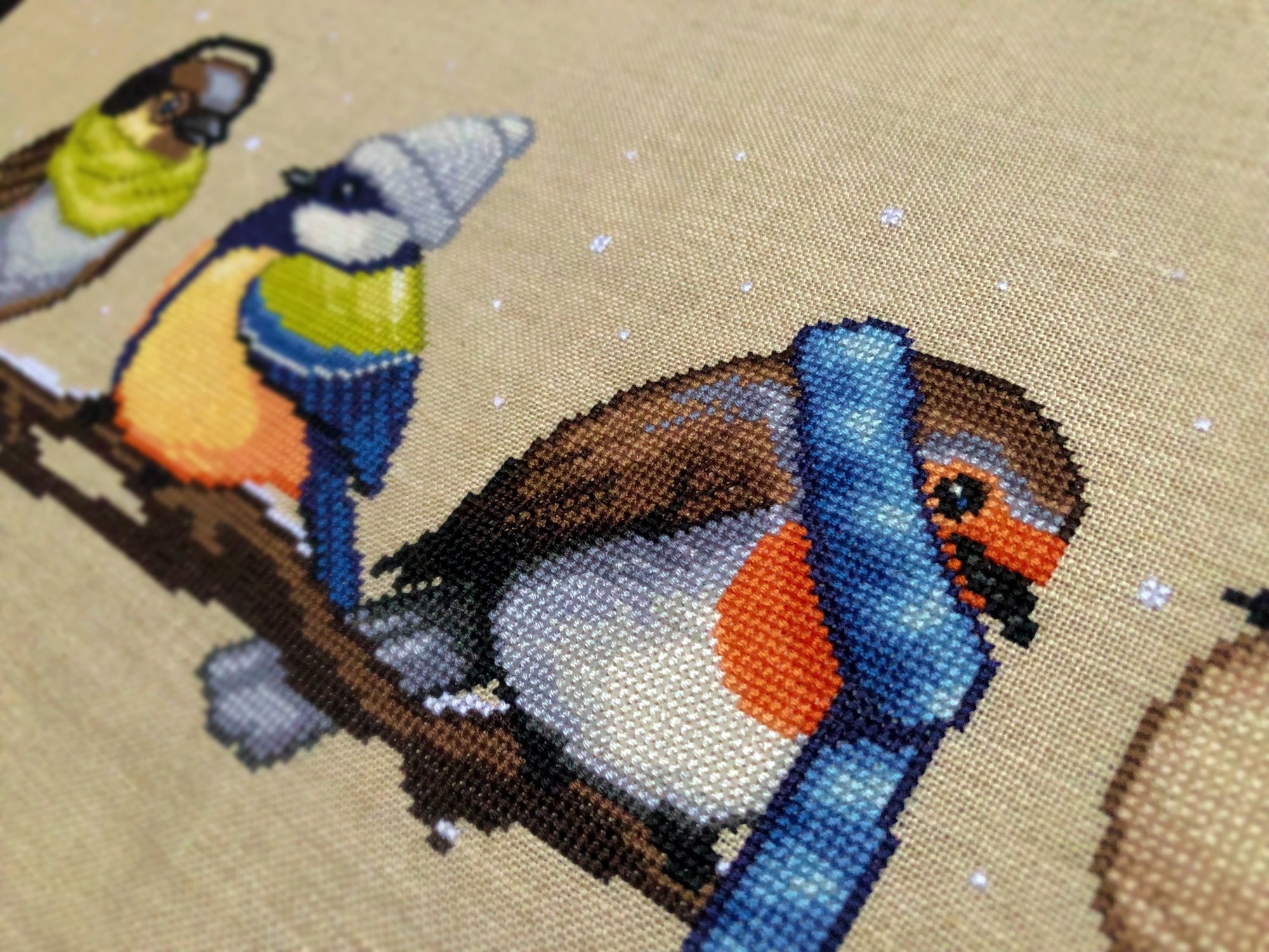 Closeup of Winter Birds cross stitch pattern. Image is zoomed in on the robin. Very cute and very round. The robin is wearing a blue, striped scarf and looks very warm. The robin is facing the wren.