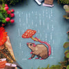 Topdown image of Rainy days rat cross stitch pattern. Fabric is turqois-blue, rat is brown with a pink tail. He is holding a mushroom over his head. His head is tilted upwards. His little paw is in the air. The mushroom keeps the rain out of his face