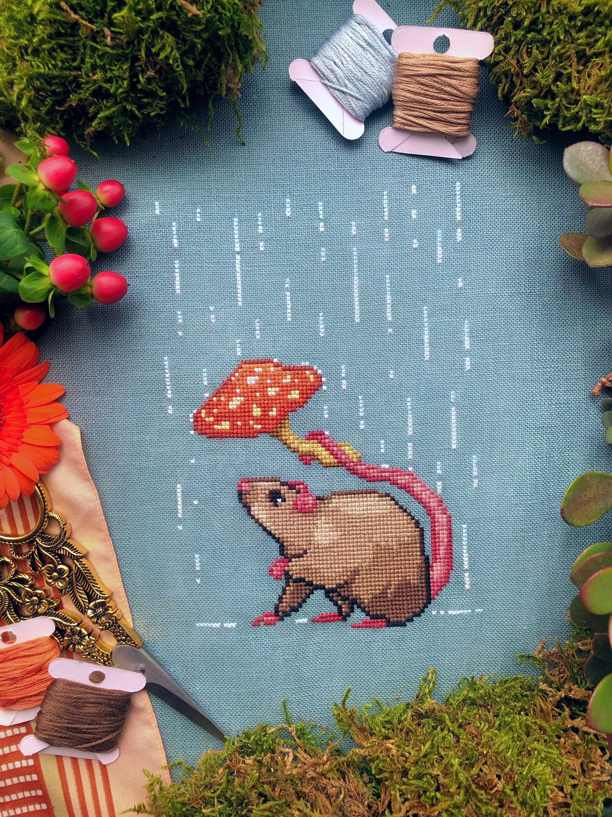 Topdown image of Rainy days rat cross stitch pattern. Fabric is turqois-blue, rat is brown with a pink tail. He is holding a mushroom over his head. His head is tilted upwards. His little paw is in the air. The mushroom keeps the rain out of his face