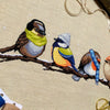 Flatlay of Winter Birds cross stitch pattern. Stitched item is surrounded by decorations. Finished piece is of medium to large size. Colors are yellow, brown, orange, blue, green, white. There are four birds sitting on a branch wearing winter gear.