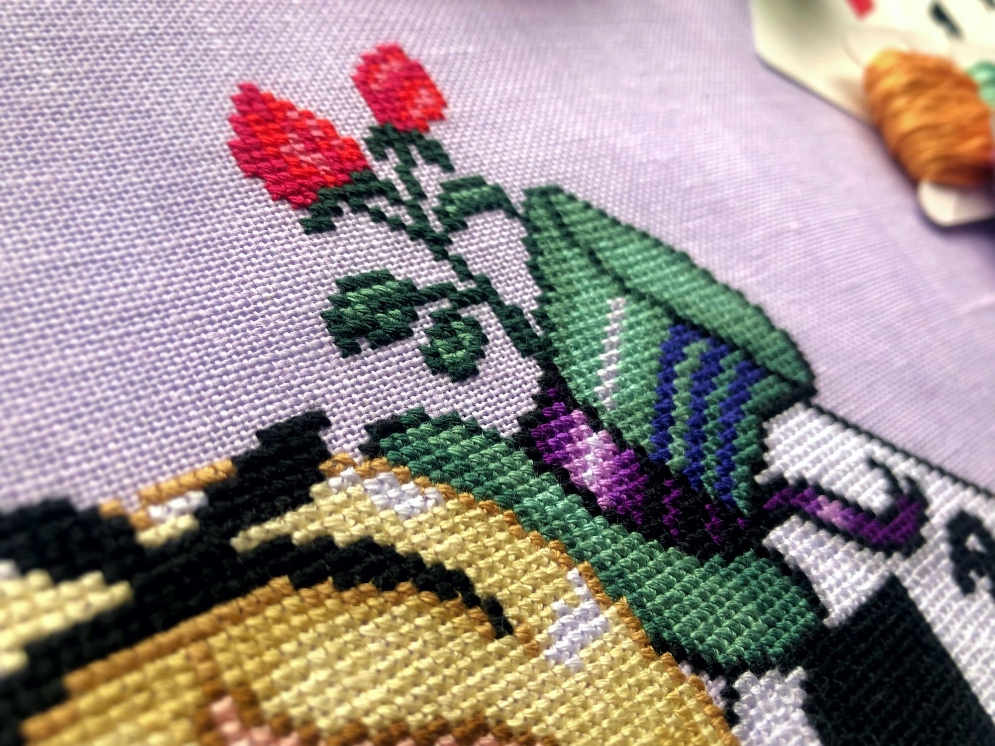 Closeup of top right side of cross stitch pattern. Here the hat of the Mad Hatter is visible. It is tall and green with blue markings, and with a purple ribbon wrapped around it. Behind it are more red roses.